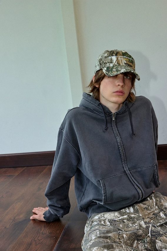 Graphite hoodie worn zipped with a camouflage cap and camouflage trousers, seated on a wooden floor