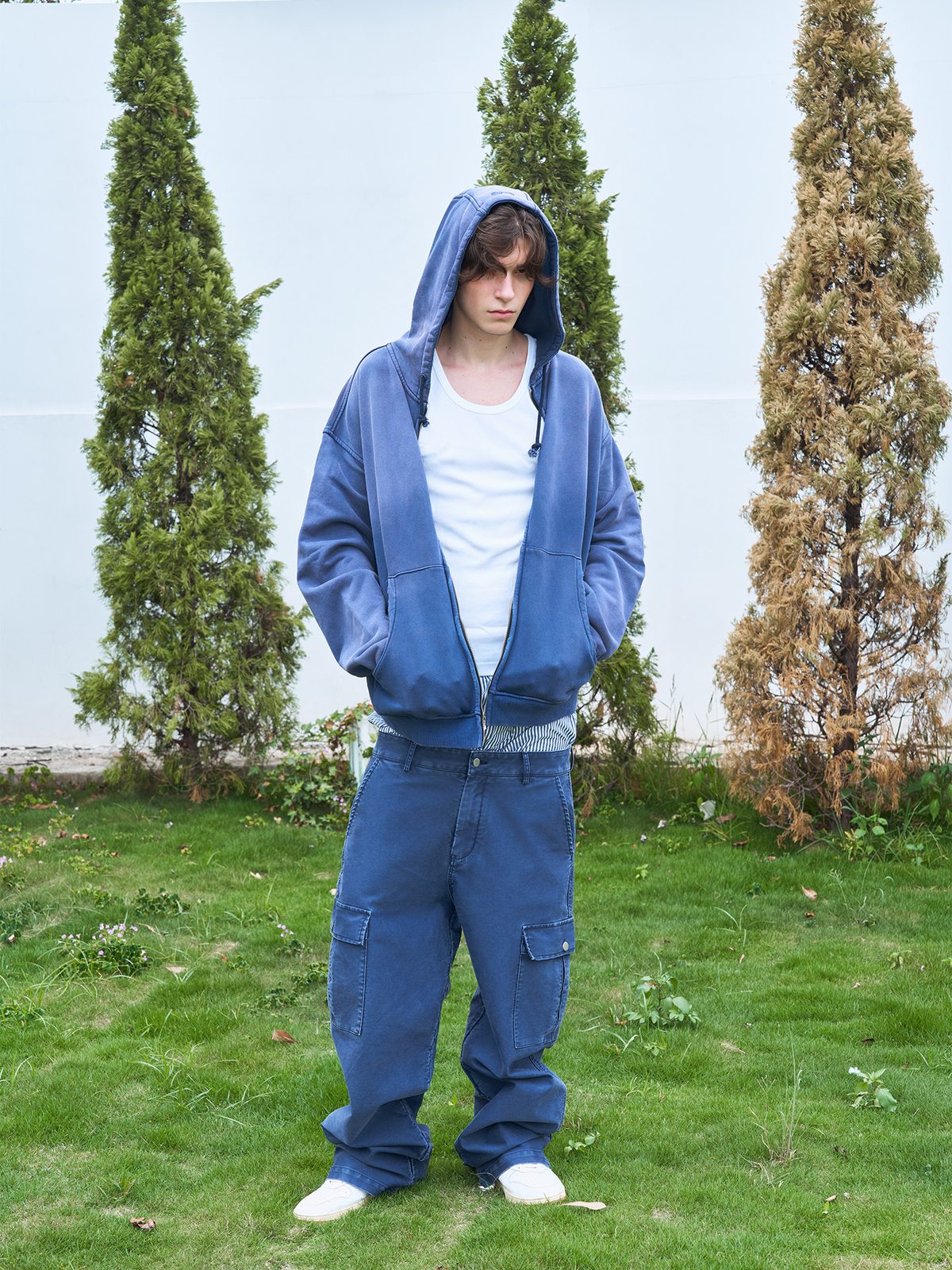 Campaign photograph of the washed indigo zip hoodie worn open over a white vest with matching washed cargo trousers, shot outdoors against a white wall and conifers