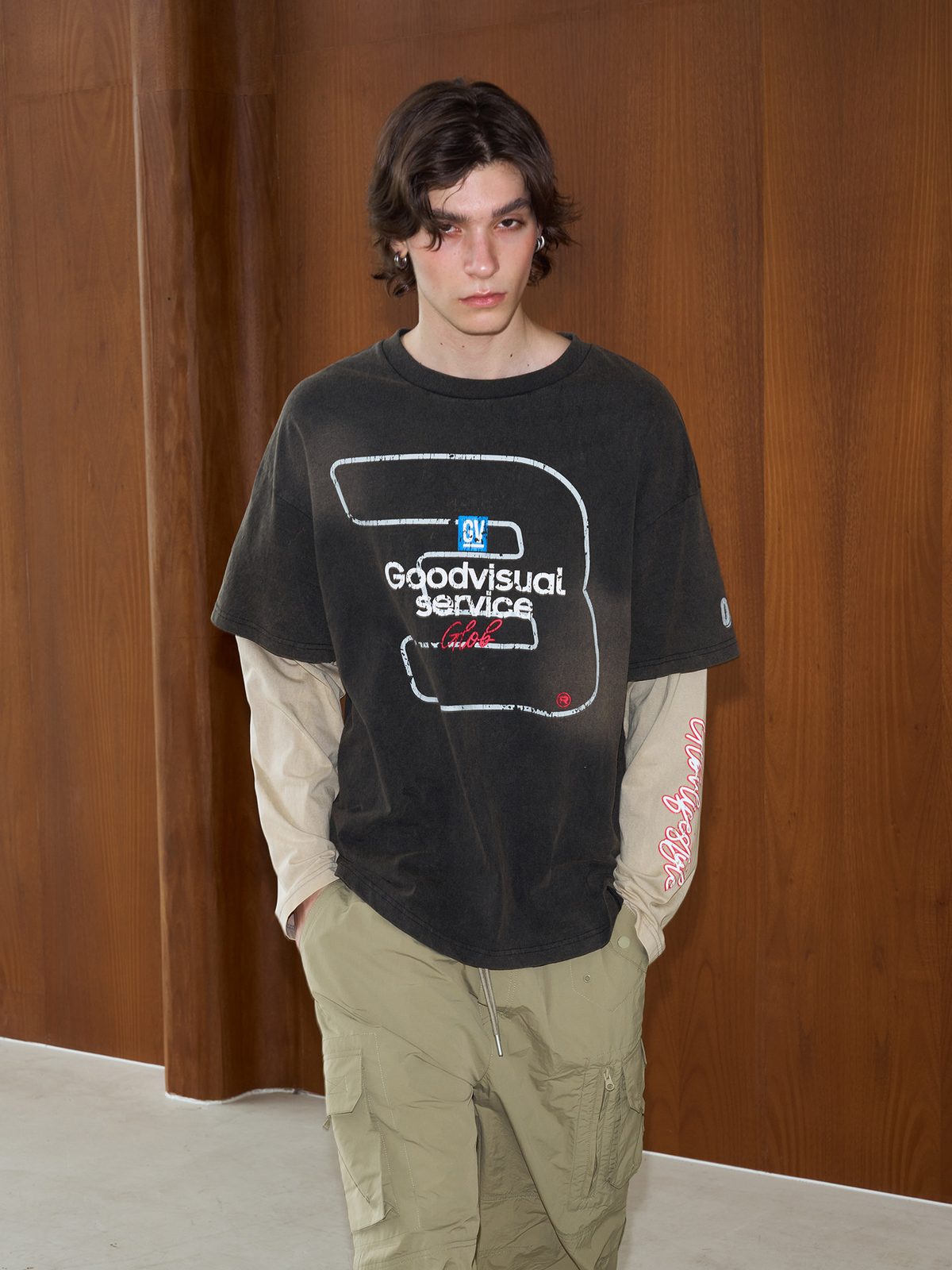 Layered-sleeve tee worn with olive trousers against a wood wall