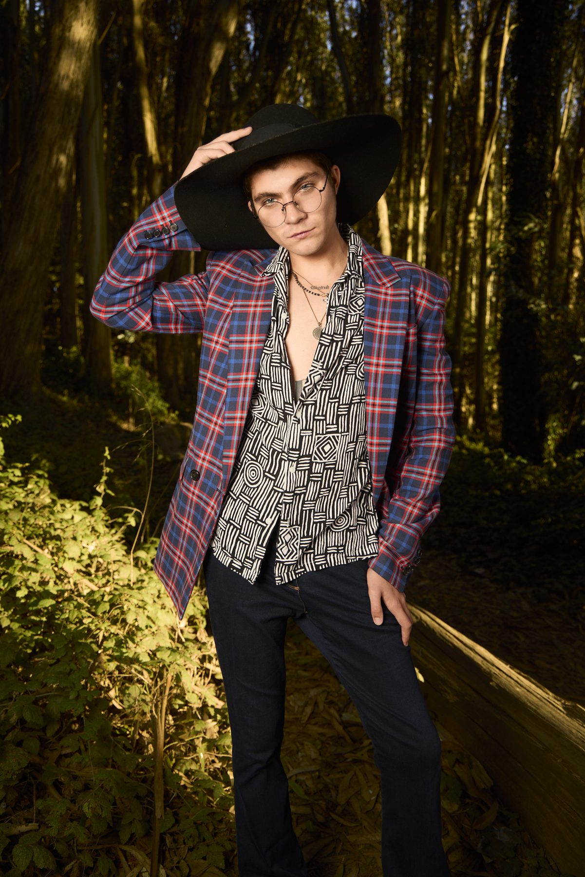 Model with a hand on the brim of a wide black hat, plaid blazer, sunlit grove