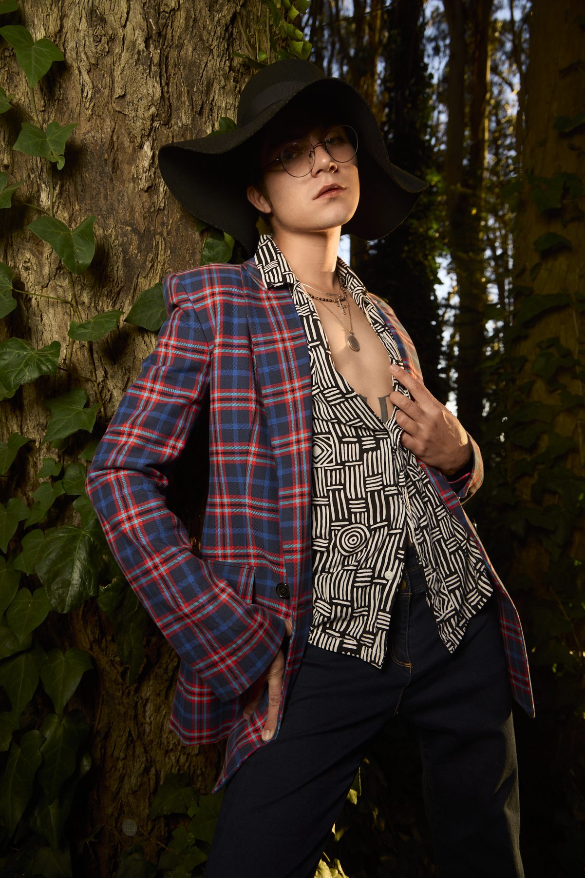 Model leaning against an ivy-covered trunk, blazer open over a printed shirt