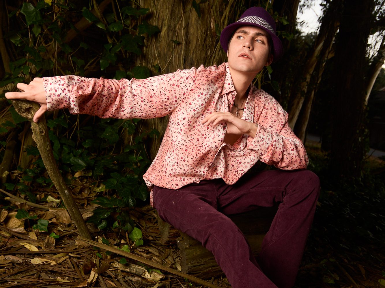 Model seated on a stump in a pink floral shirt and purple fedora, hard flash at dusk