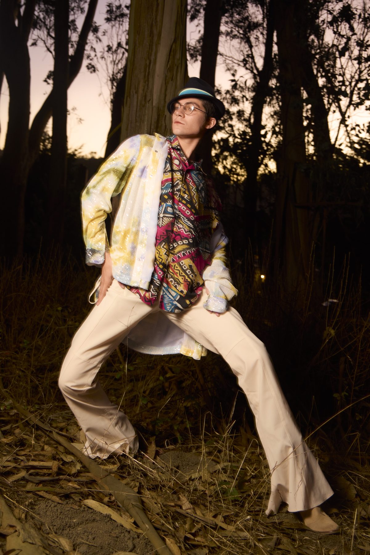 Model in a sequinned jacket over a printed shirt, wide stance, night flash